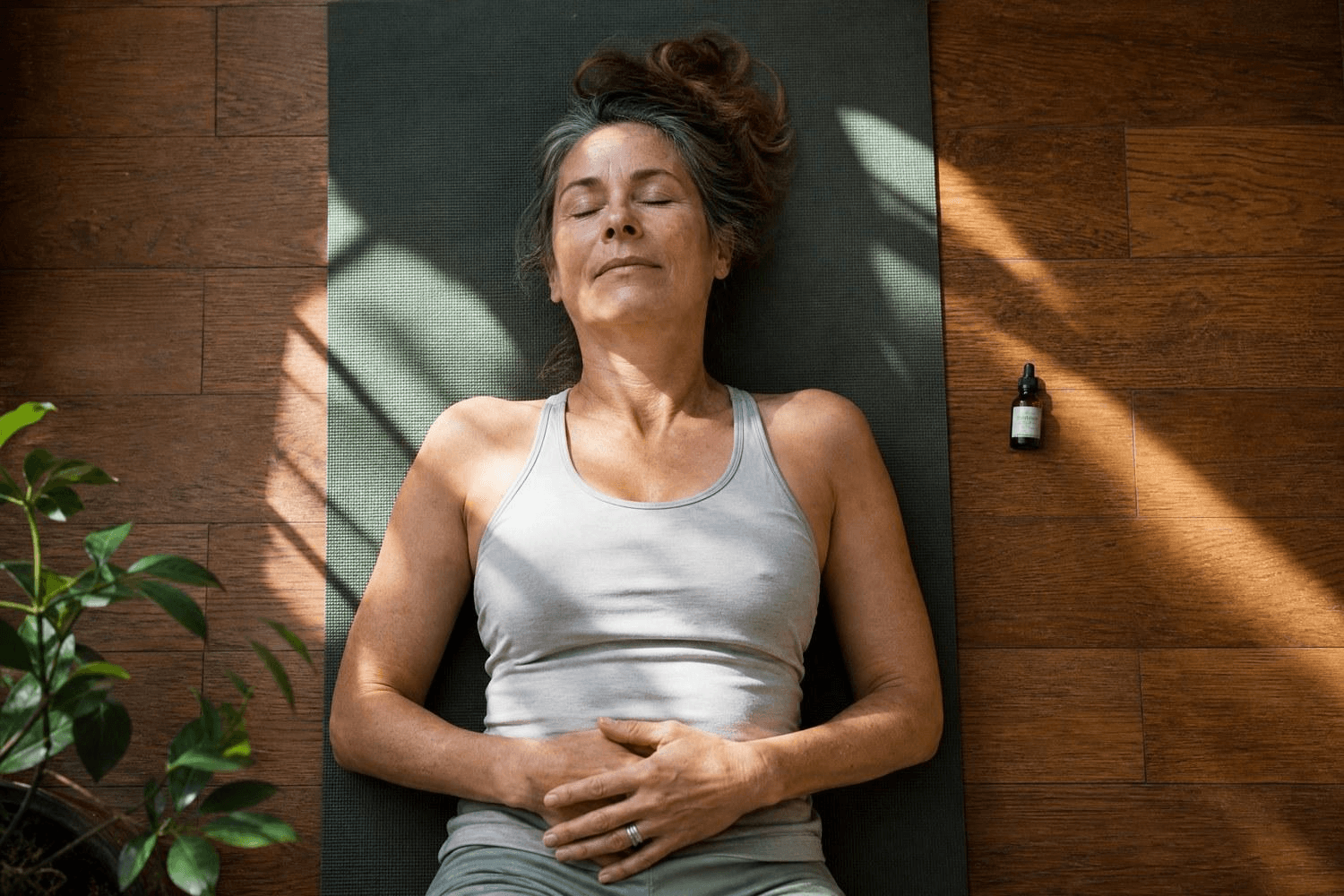 Eine Frau mittleren Alters liegt entspannt auf einer Yogamatte, Augen geschlossen, neben ihr ein kleines CBD-Öl-Fläschchen – Vogelperspektive.