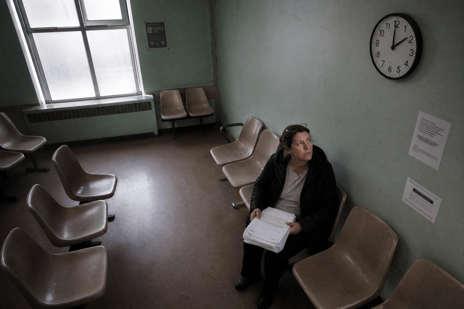 Eine mittelalte Frau sitzt allein in einem leeren Wartezimmer und blickt auf eine Uhr an der Wand – sie hält einen Stapel medizinischer Unterlagen auf dem Schoß.