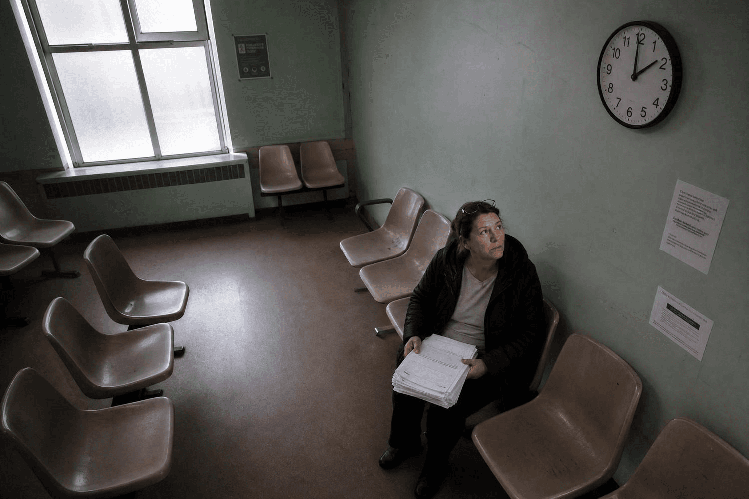 Eine mittelalte Frau sitzt allein in einem leeren Wartezimmer und blickt auf eine Uhr an der Wand – sie hält einen Stapel medizinischer Unterlagen auf dem Schoß.