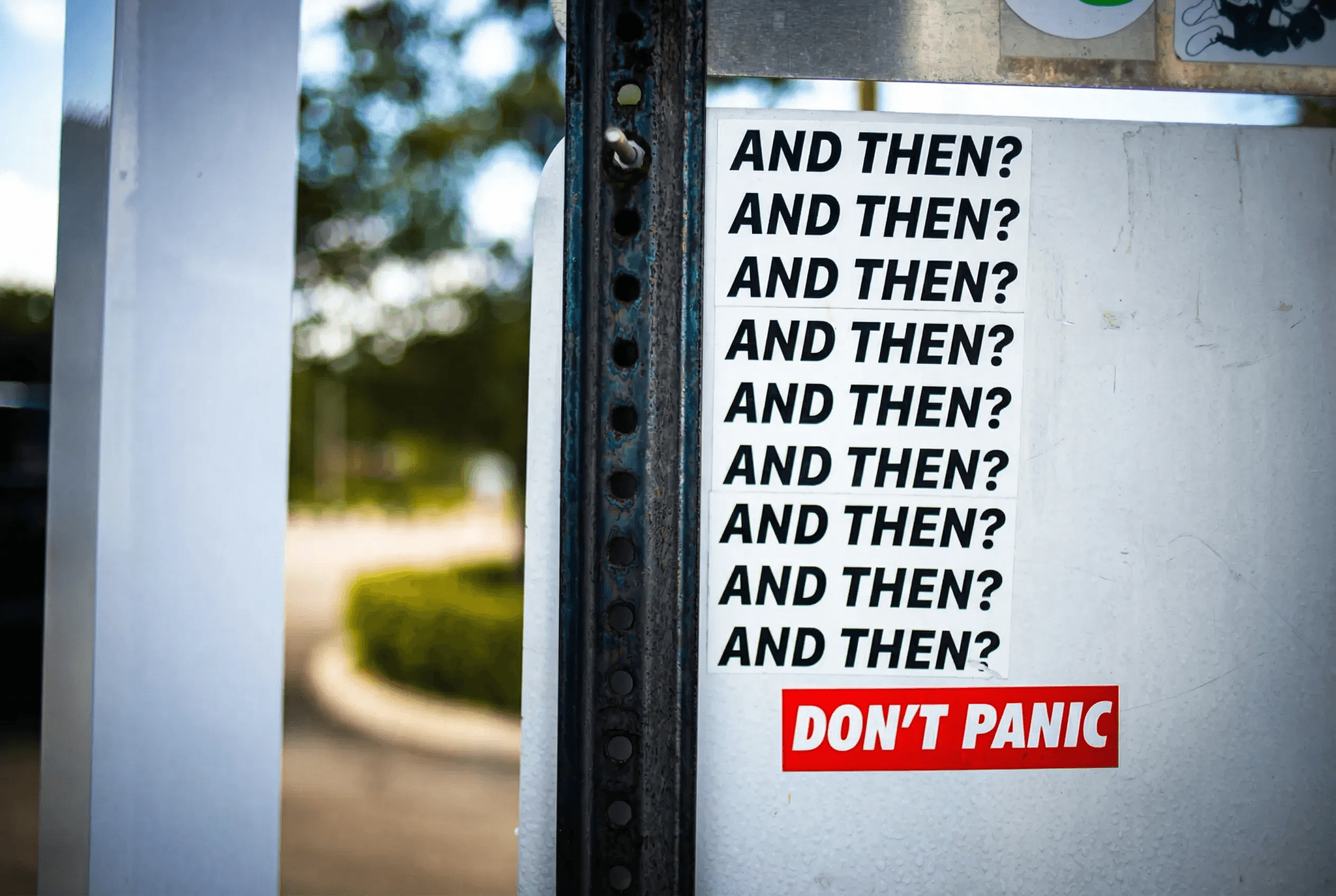Streetart-Poster mit wiederholtem 'AND THEN?' Text und rotem 'DON'T PANIC' Banner an Metallpfosten