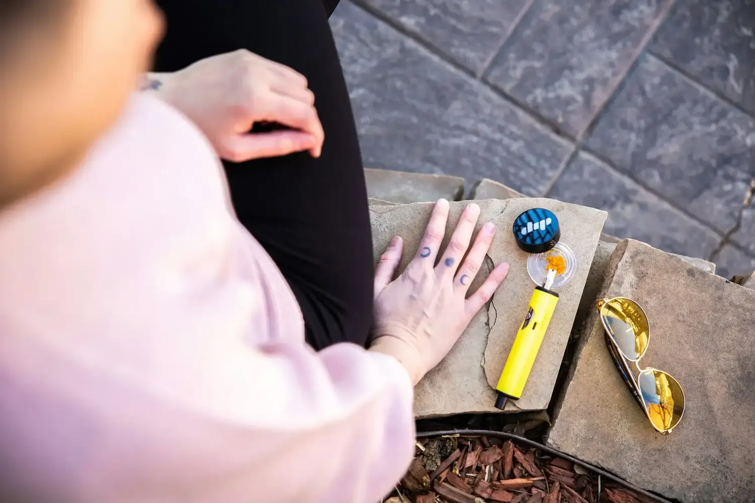 Hand mit Mondtattoos neben gelbem Dab-Pen, Konzentrat und goldener Sonnenbrille