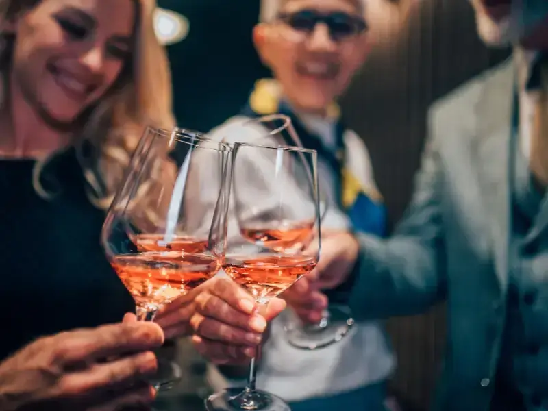 Hände stoßen mit roséfarbenen Weingläsern an, Gäste unscharf im Hintergrund