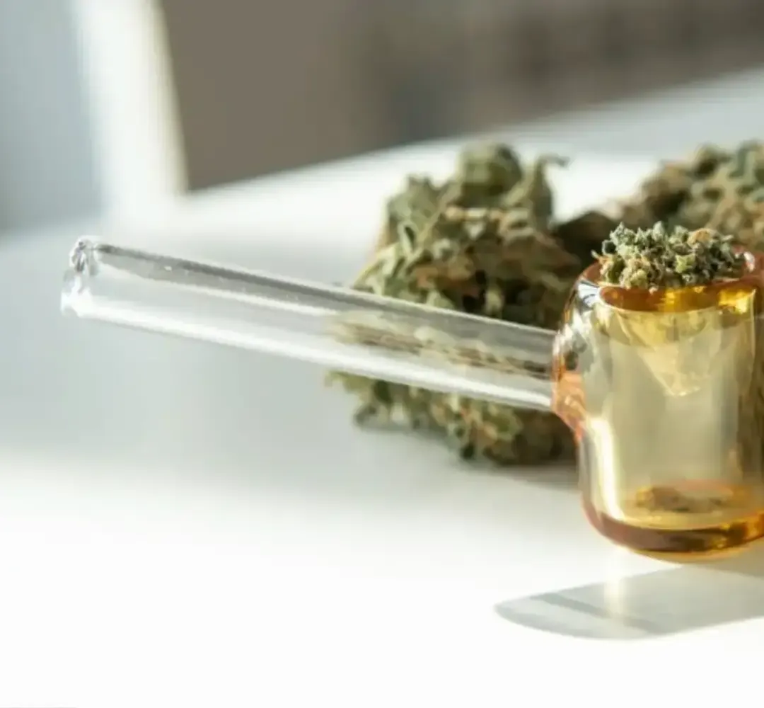 Klare Glas-Pfeife neben Cannabisblüten auf weißer Fläche