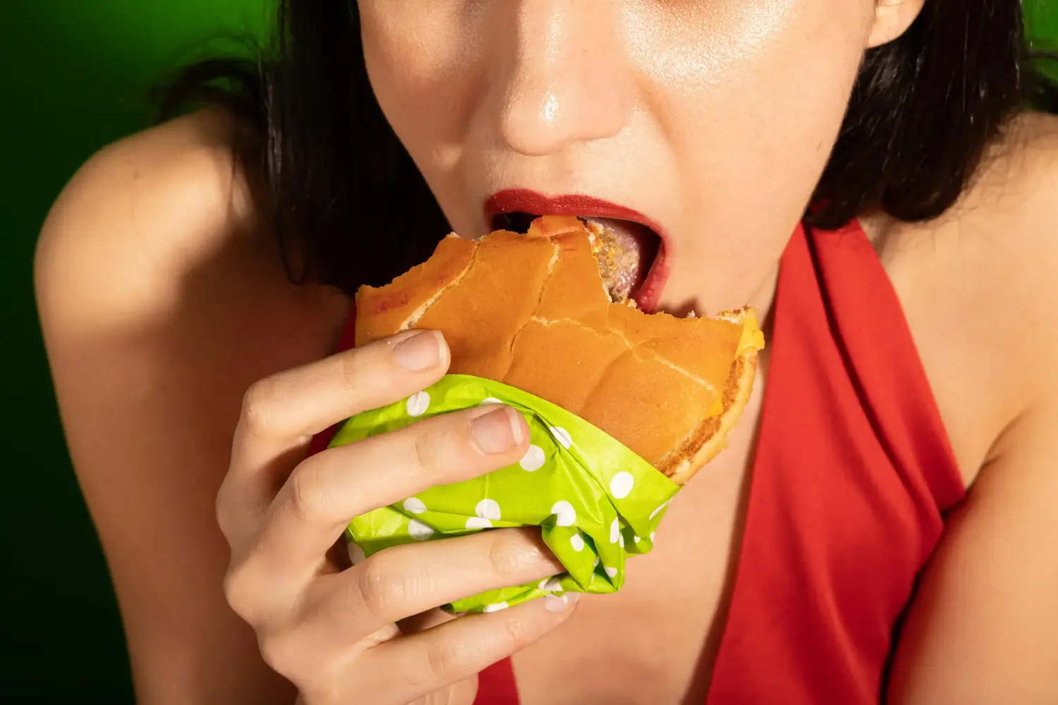 Nahaufnahme: Person mit rotem Top beißt in Burger mit grünem Tupfenpapier