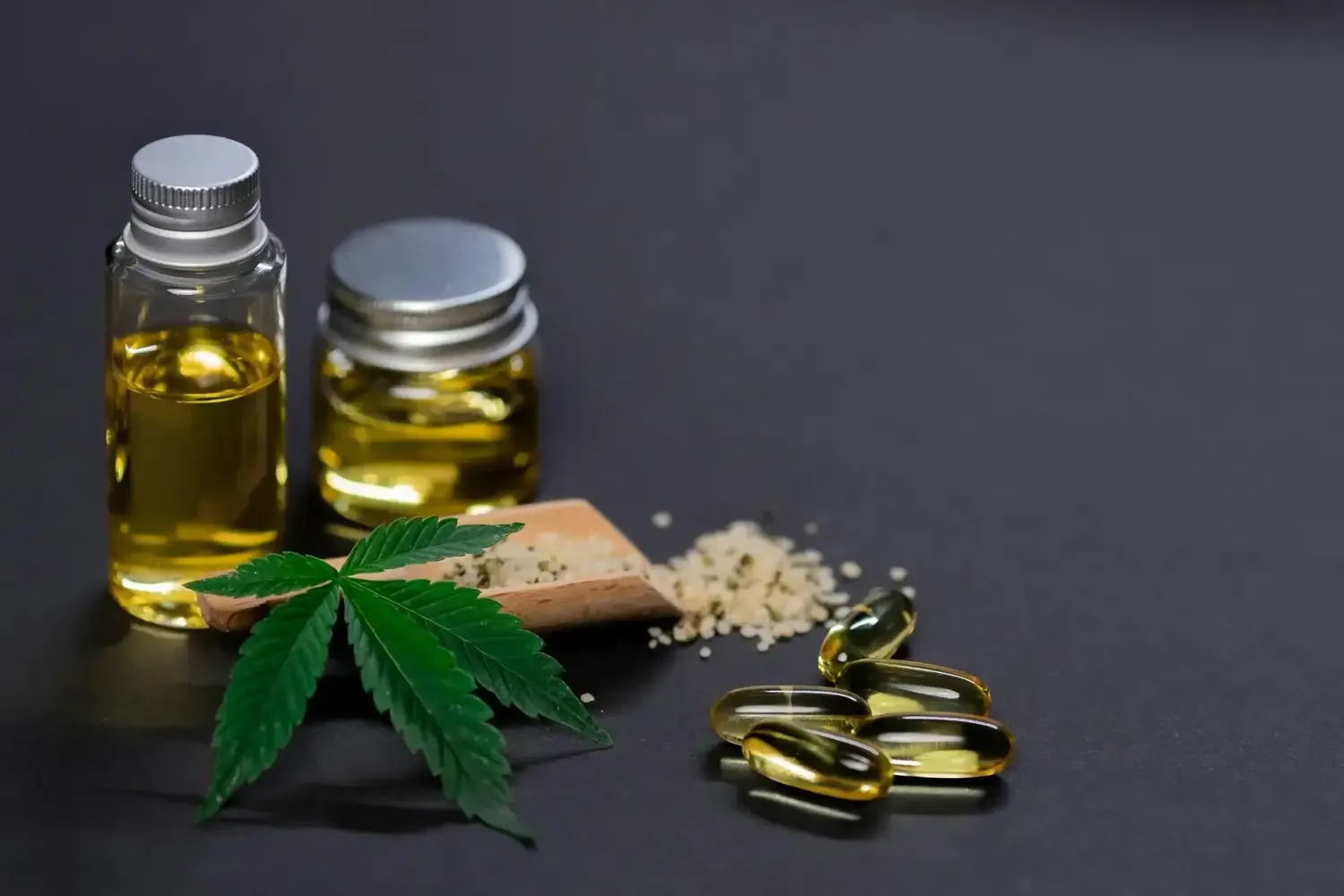 Zwei Glasflaschen mit goldenem Öl, Gelkapseln, Hanfblatt und Granulat auf dunklem Untergrund