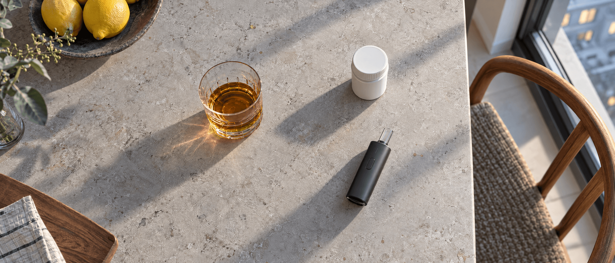 Heller Tisch am Fenster mit Glas Alkohol, weißem Arzneimittelbehältnis und schwarzem Vaporizer.