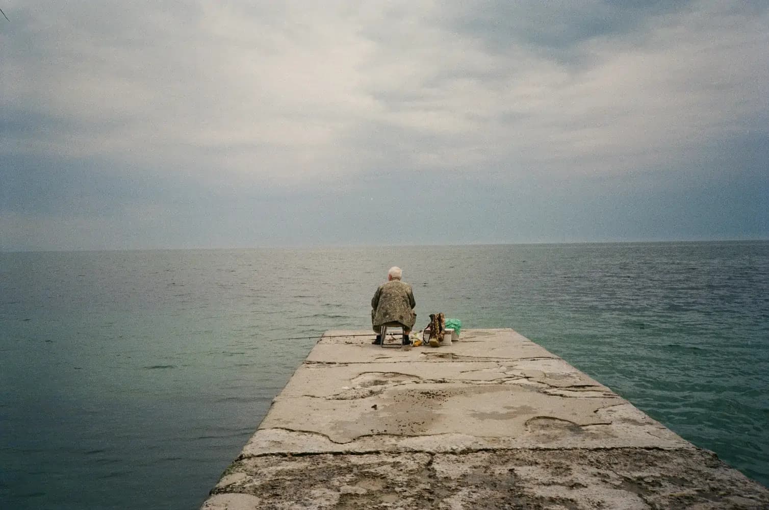 Ältere Person auf Stuhl am Ende eines Betonstegs, mit Angelausrüstung, Blick aufs Meer