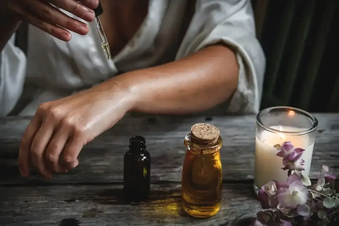 Person träufelt CBD-Öl mit Pipette auf Innenseite des Unterarms, Flaschen und Kerze daneben