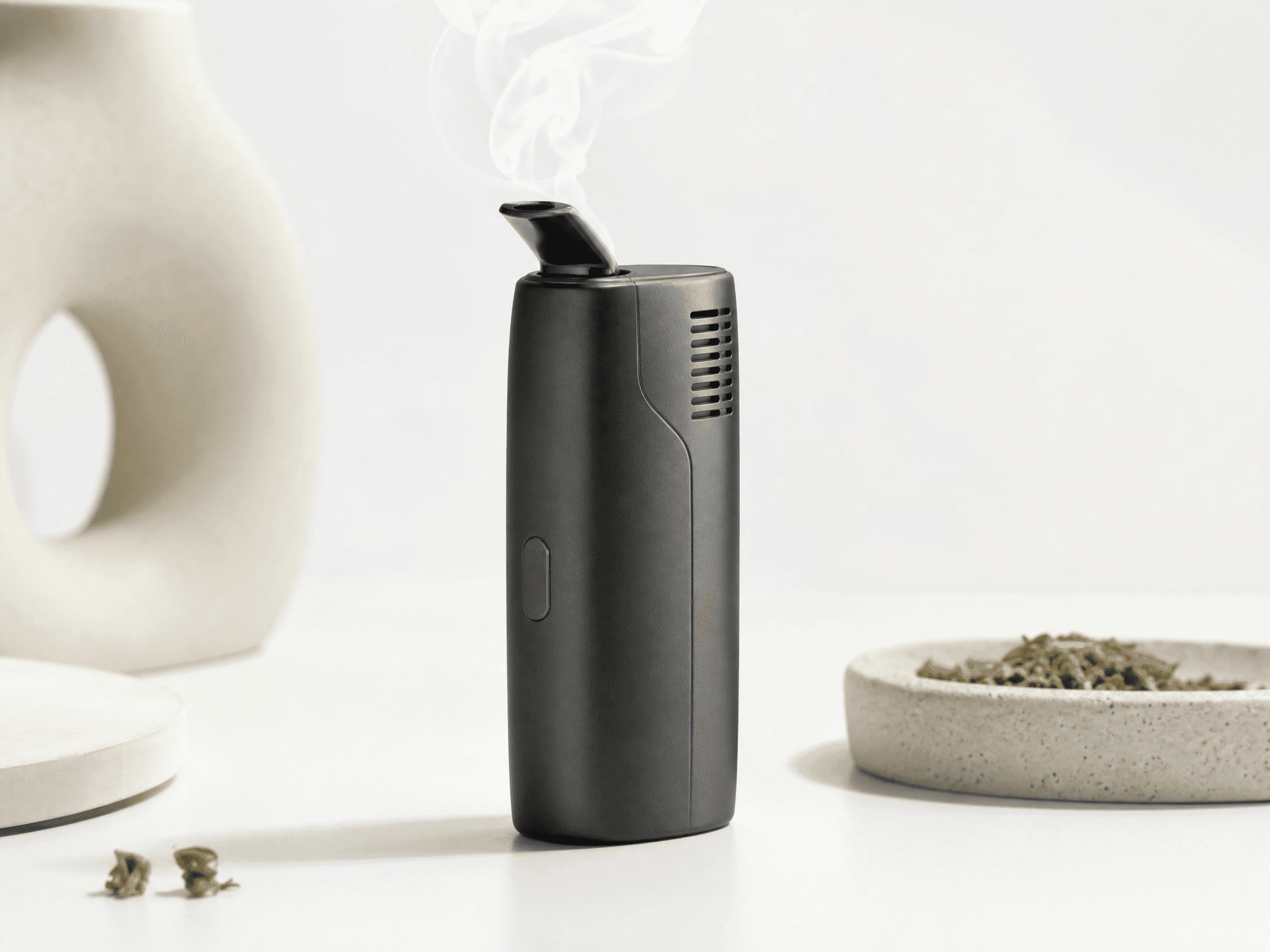 Kompakter schwarzer Trockenblüten-Vaporisator auf weißer Oberfläche, aus dessen Mundstück feiner Dampf aufsteigt – daneben getrocknete Cannabisblüten auf einem Steinteller