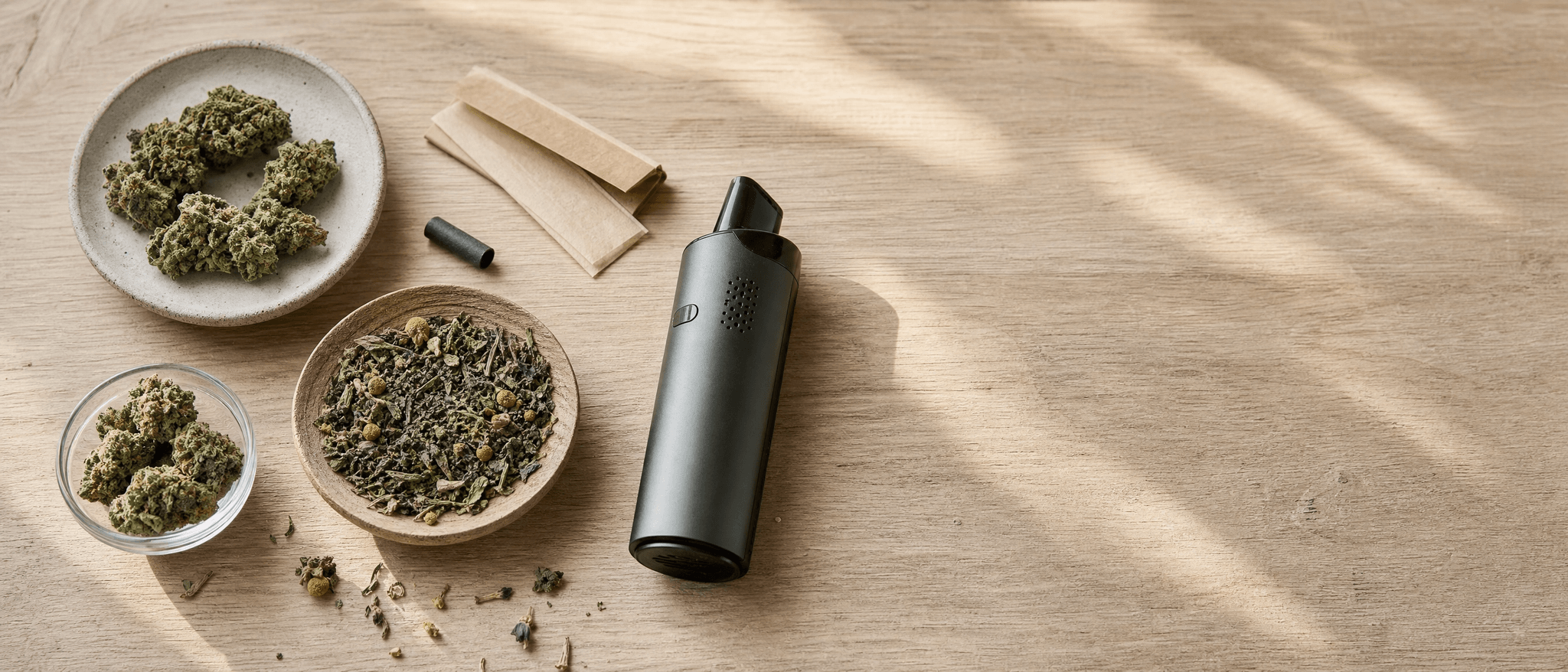 Vogelperspektive auf Cannabisblüten, getrocknete Kräuter, ungebleichte Papers und einen Blütenvaporizer auf hellem Holzti