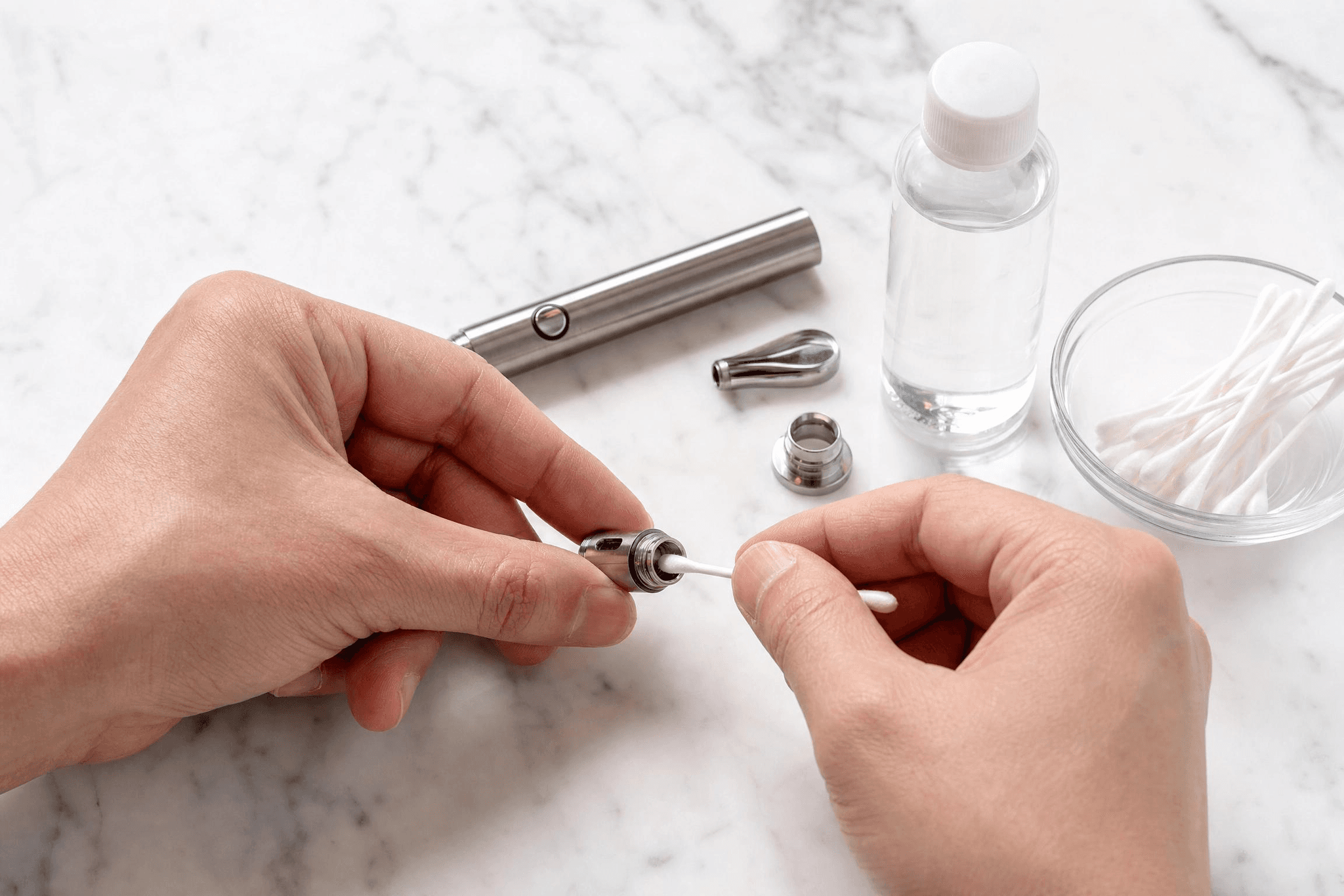 Hände reinigen das Heizelement eines zerlegten Wax Pens mit einem Wattestäbchen, daneben Isopropanol-Flasche und Wattestäbchen-Schale auf Marmoruntergrund.