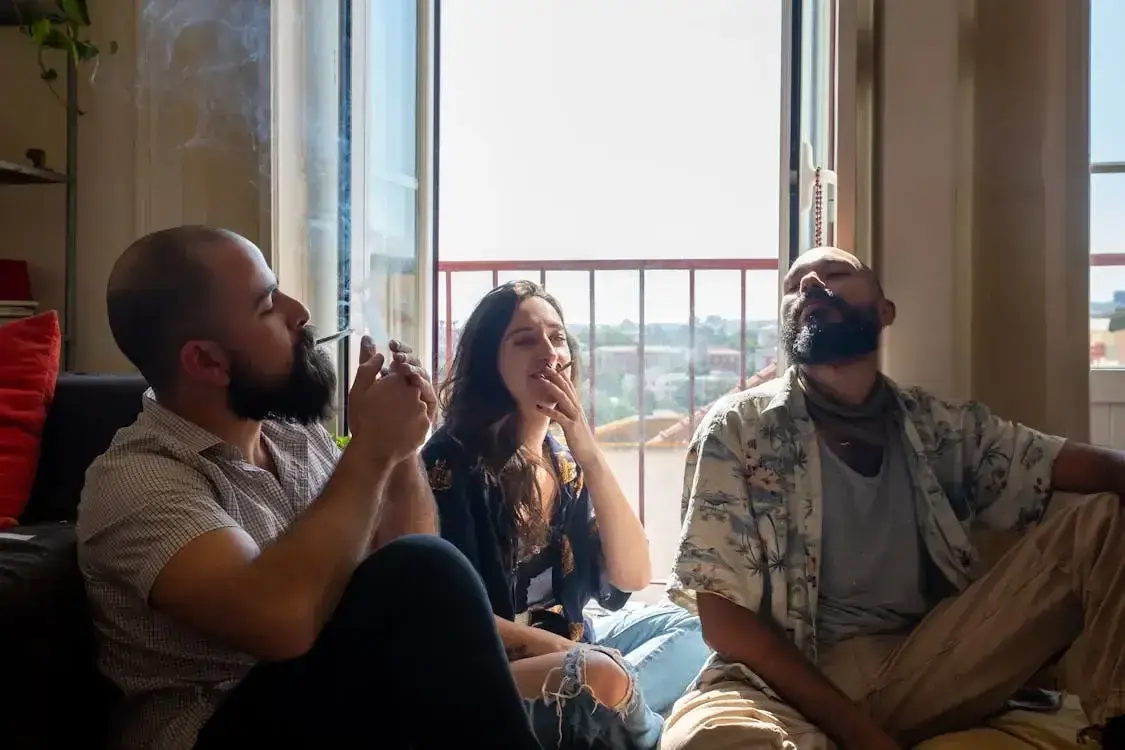 Drei junge Erwachsene sitzen am offenen Fenster und rauchen Joints.