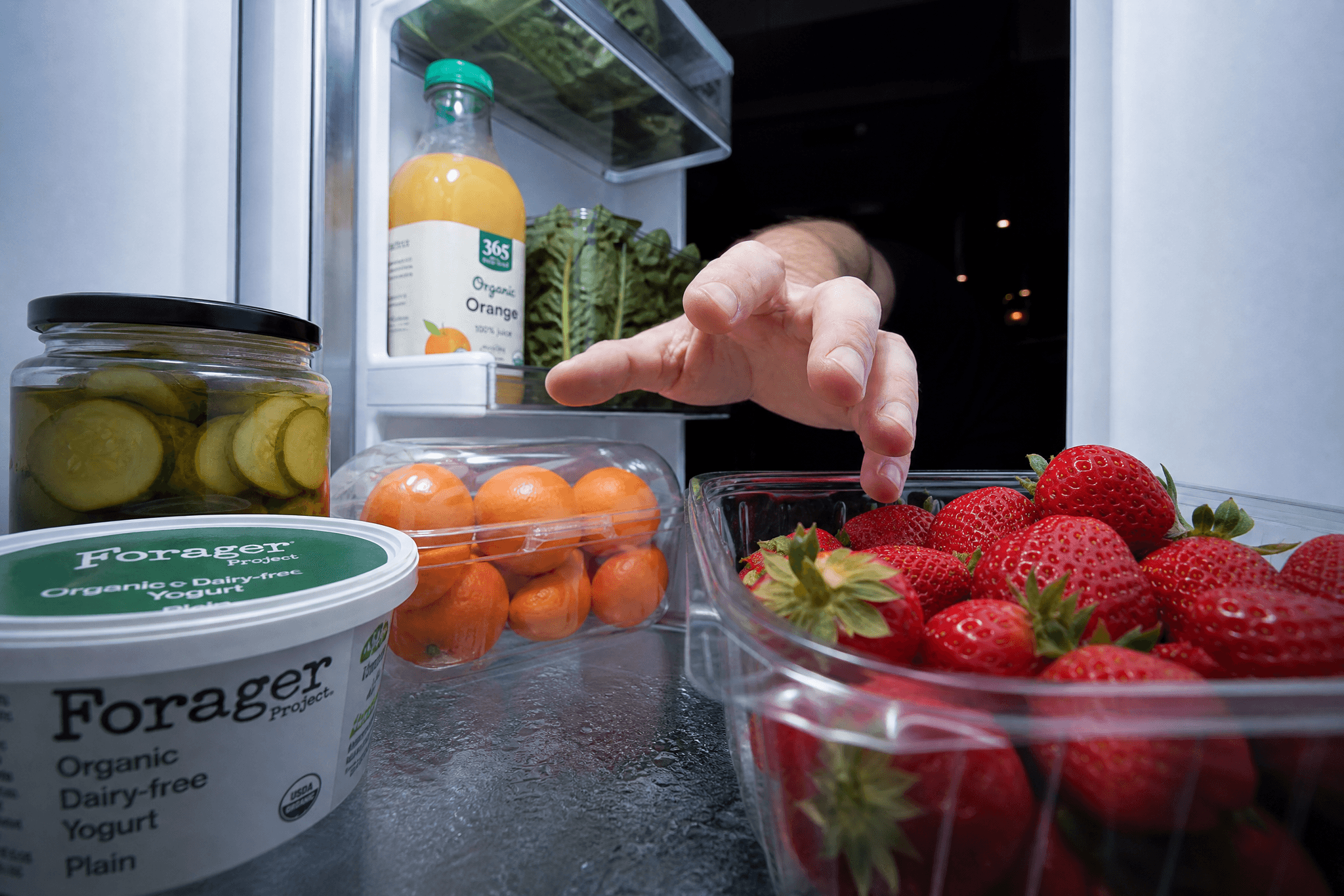Perspektive aus dem Inneren eines geöffneten Kühlschranks: Eine Hand greift von hinten nach frischen Erdbeeren in einer Glasschale; im Vordergrund sind außerdem Kirschtomaten, ein Glas Essiggurken, eine Flasche Orangensaft sowie ein Becher veganer Joghurt zu sehen – nächtliche Küchenszene, die den möglichen Appetit-steigernden Effekt von Cannabis illustriert.