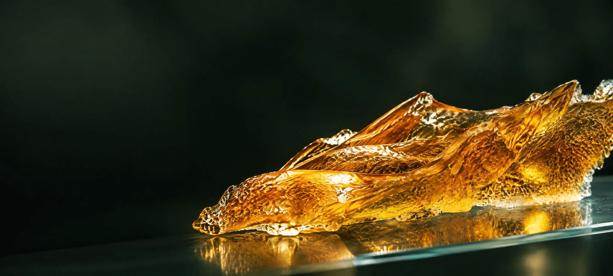 Makroaufnahme von goldfarbenem, transparentem Rosin-Konzentrat auf einer spiegelnden Glasfläche vor dunkelgrün-schwarzem Hintergrund.