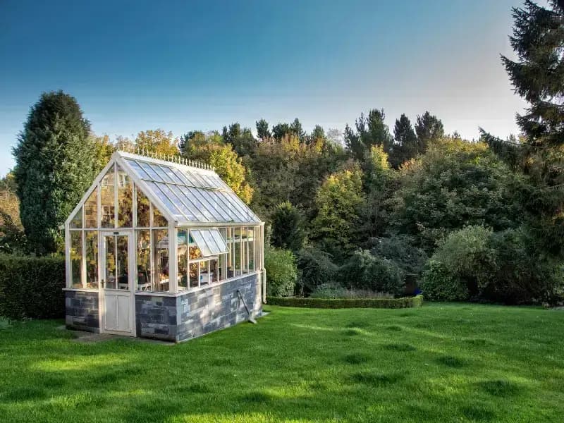 Weißes Glasgewächshaus auf grünem Rasen, umgeben von Bäumen und Hecken