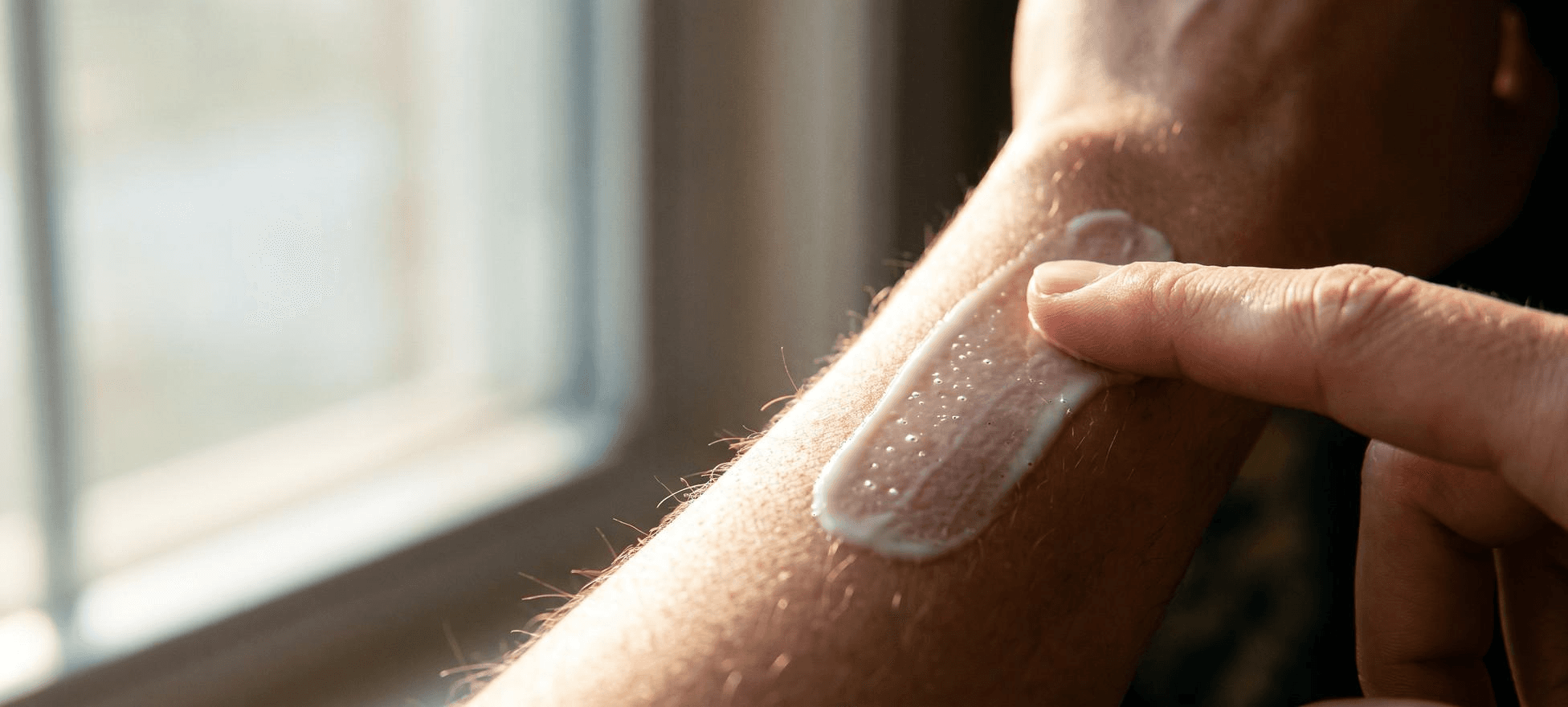 Makroaufnahme: Ein Finger verteilt eine dünne Creme auf der Haut eines Unterarms, Streiflicht hebt feine Härchen hervor.