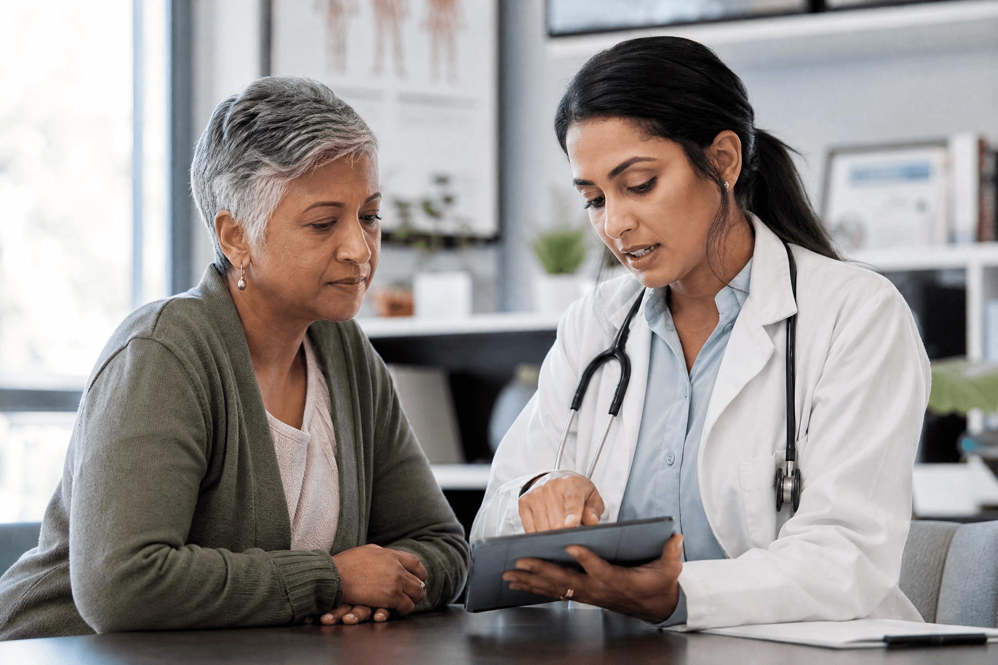 Ärztin im weißen Kittel zeigt einer älteren Patientin etwas auf einem Tablet während eines Beratungsgesprächs in einer Arztpraxis.