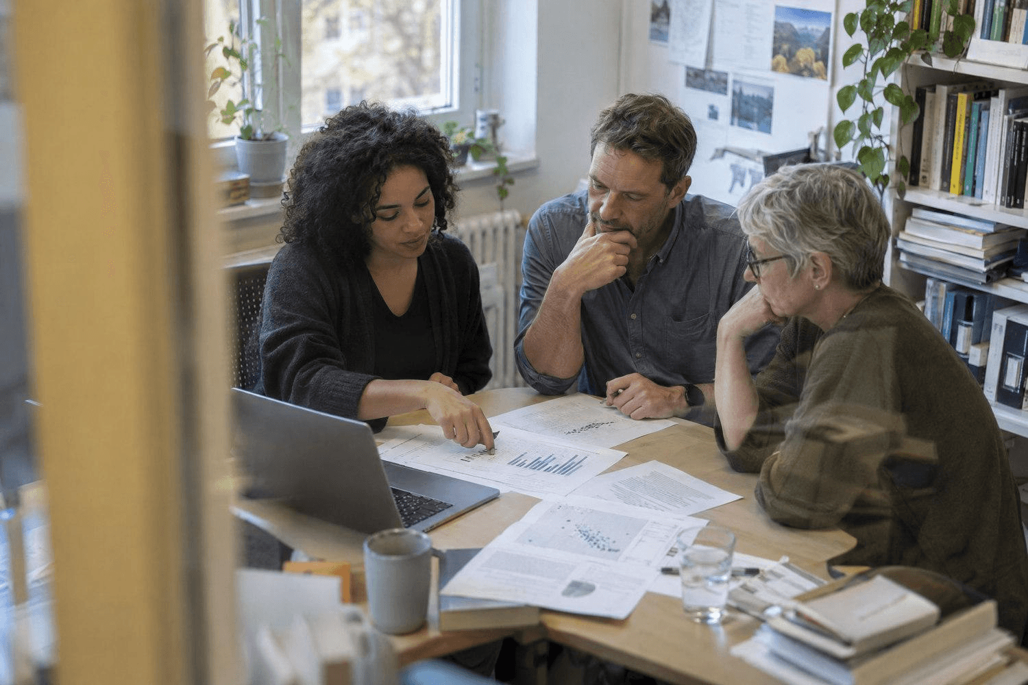 Drei Erwachsene besprechen an einem Tisch wissenschaftliche Ausdrucke, Diagramme und Daten auf einem Laptop.